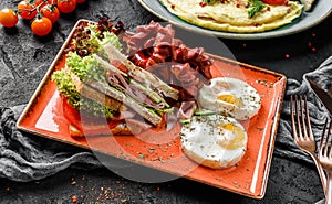 Delicious breakfast with fried eggs, sandwiches with ham and vegetables, and sausages served on plate over dark background.