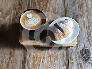 Delicious Breakfast; Heart love Latte art coffee in Black cup and Croissant topped with icing sugar
