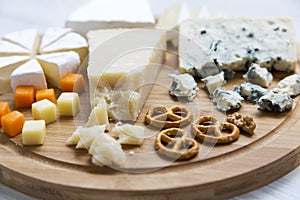 Delicious cheeses: camembert, blue cheese, brie, parmesan, mimolette and edam with pretzels and walnuts on round wooden board.