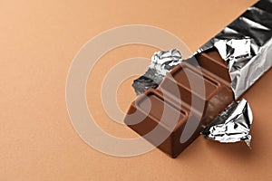 Delicious chocolate bar wrapped in foil on light brown background, closeup. Space for text