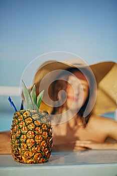 Delicious cocktail in pineapple. Summer vacation. Beautiful young woman in hat relaxing in pool with drink. Girl enjoying warm