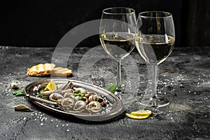 Delicious cooked sea escargo snails with herbs, butter, garlic on metal plate with forks. wine glass. gourmet food. Restaurant