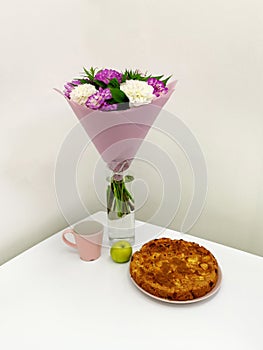 Delicious dessert apple pie on a plate, a bouquet of flowers and a pink mug on a white table. Cozy table setting for