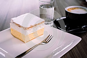 Delicious desset cream cake on the white square plate served with cappuccino and water at Bled lake, Slovenia