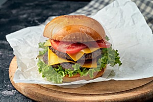 Delicious double burger with beef, tomatoes, lettuce, melted cheese on white parchment. Dark background. American fast food