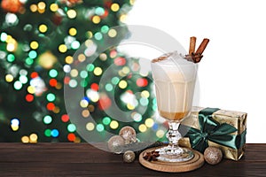 Delicious eggnog in glass, gift box, festive ornaments on wooden table against Christmas tree, space for text