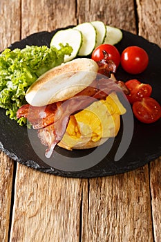 Delicious English muffin sandwich with scrambled eggs, bacon, cheese and vegetables close-up on a slate board. vertical