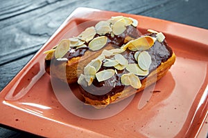 Delicious French dessert - custard eclairs, chocolate icing and almonds in a red plate. Black wood background