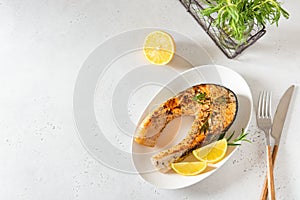 Delicious fried salmon steak with rosemary, lemon and pepper served on a white plate on white table. Top view, copy space.