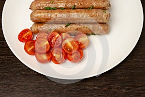 Delicious fried tasty hot sausages with cherry tomatoes on white ceramic plate and wooden background above view