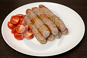 Delicious fried tasty hot sausages with cherry tomatoes on white ceramic plate and wooden table background