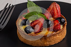 Delicious fruit cake or tartlet with fresh strawberries, cranberries and mint leaf on a black plate and a fork next to it