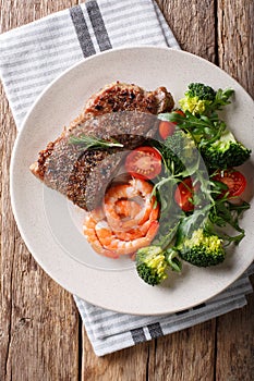 Delicious grilled beef steak with prawns and broccoli, tomatoes, arugula closeup on a plate. Surf and Turf. Vertical top view