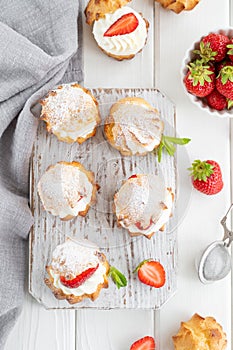 Delicious homemade small cakes profiterole choux pastry with custard, strawberry and icing powder on the white wooden background.