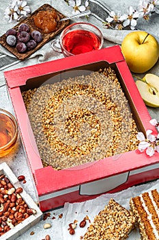 Delicious honey biscuit cake with cream, apples, cherries and peanuts in paper box for delivery on light background with cups of