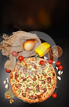 Delicious Italian pizza on a black burlap table with food ingredients: cherry tomatoes, yellow bell peppers, onions, garlic,