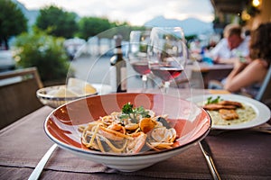 Delicious italian seafood pasta in red plate with two glasses of red wine in outdoor restaurant in Bellagion town in Italy