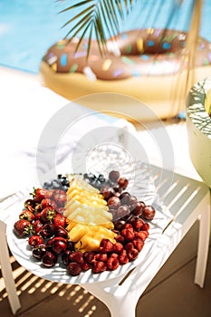 Delicious juicy fruit and berry set on the table by the pool. Pineapple, cherries, strawberries, raspberries and grapes on a plate