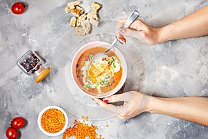 Delicious lentils soup with heavy cream on concrete table with red bell pepper, bread toasts. Female hands holds spoon.