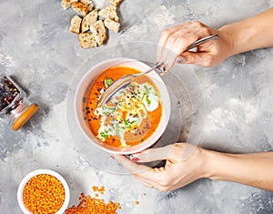 Delicious lentils soup with heavy cream on concrete table with red bell pepper, bread toasts. Female hands holds spoon.