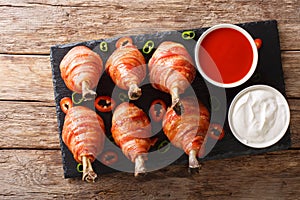 Delicious Lollipops chicken legs wrapped in bacon with sauces close-up on a slate board. horizontal top view
