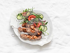 Delicious lunch, appetizer, tapas - chicken kebabs and fresh vegetable cucumbers tomatoes salad on a light background, top view