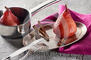 Delicious pear in wine with cinnamon and anise on silver plate and in stewpot on grey concrete surface with pink napkin, knife and