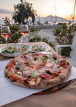 Delicious pizza with cheese, bacon, salami, mushrooms and pepper and two glasses of wine against background of port in Greece