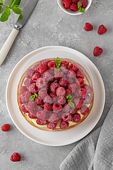 Delicious raspberry tart with vanilla custard cream sprinkled with powdered sugar on a white plate on a gray concrete background.