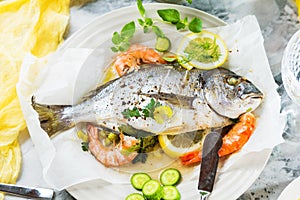 Delicious roasted dorado or sea bream fish with lemon and fresh Prawns , fresh parsley and spinach on white platter .