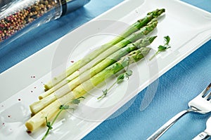 Delicious side dish for main courses - boiled asparagus with microgreen in a white plate on a blue tablecloth