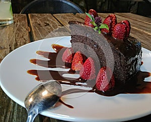 Delicious slice of rich dark chocolate cake with chocolate icing and fresh red strawberries. On wooden table.