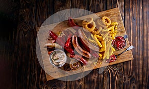 Delicious snacks with grilled sausages, fried potato, onion rings and two glasses of beer on wooden board in rustic