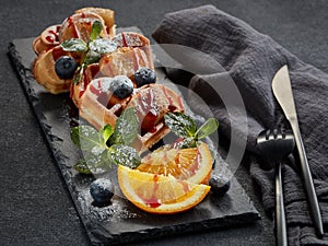 Delicious waffles heart shape with caramelized banana and berries on serving board on grey background. Dessert. Serving food