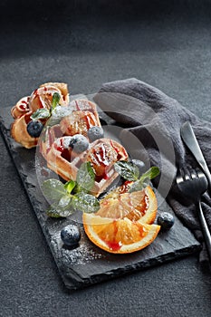 Delicious waffles heart shape with caramelized banana and berries on serving board on grey background. Dessert. Serving food