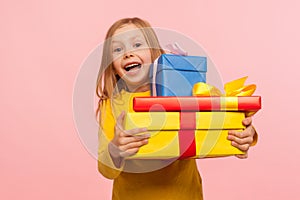 Delighted amazed little girl embracing lot of gift boxes, looking at camera with surprise and sincere childish happiness photo