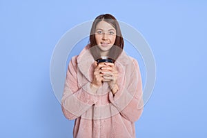 Delighted attractive woman standing isolated over blue background, looks at camera, holding disposable mug in both hands, wearing