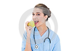 Delighted brown haired nurse in blue scrubs eating a green apple