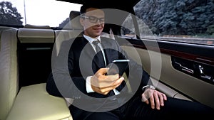 Delighted businessman riding on backseat of expensive car, using phone app