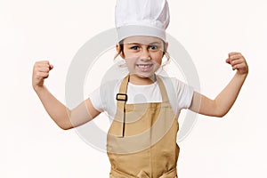 Overjoyed baby girl dressed as confectioner in chef's hat and apron clenches fists, looks at camera, on white