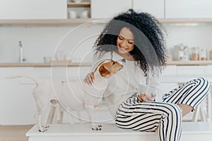 Delighted curly woman with cheerful expression poses with jack russell terrier dog at home, drinks aromatic beverage, dressed in