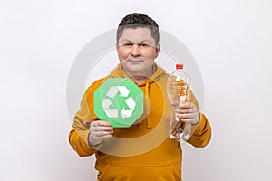 Delighted dark haired man with smile holding empty plastic bottle and green recycling sign.