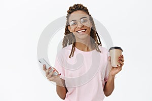 Delighted happy and amused charming african american girl with dreads in glasses holding paper cup of coffee and