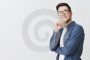 Delighted happy and satisfied handsome young male student in glasses and blue shirt holding hand on chin and smiling