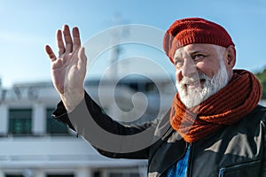 Delighted pensioner waving goodbye. White bearded man in red knitted hat and scarf