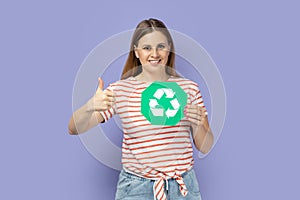 Delighted positive woman holding green recycling sign and showing thumb up, approved gesture.