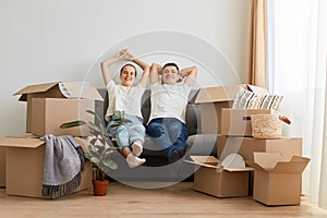 Delighted relaxed young couple sitting on sofa surrounded with carton boxes, family during moving to a new house, being happy to