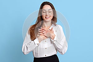 Delighted smiling employee receives praise from her college, being flattered deeply, cross hands on her chest, closes eyes.