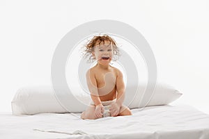 Delighted toddler, little girl with wet hair in diaper smiling while seated on soft bedspread against white studio