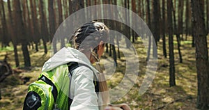 Delighted young blonde girl with braids smiling wearing backpack holding woman's hand heading through wood hikking